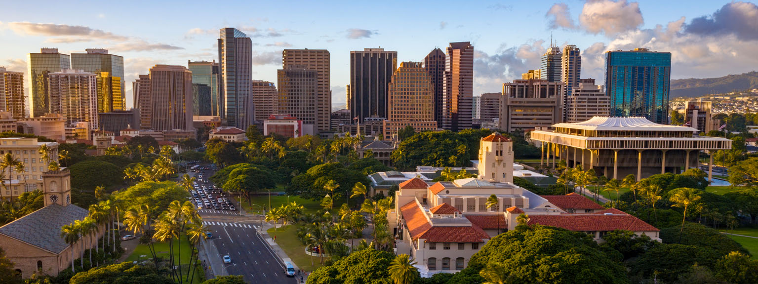 Aerial view of downtown Honolulu at sunset, featuring Honolulu Hale, the Hawai'i State Capitol, and the city skyline surrounded by palm trees.